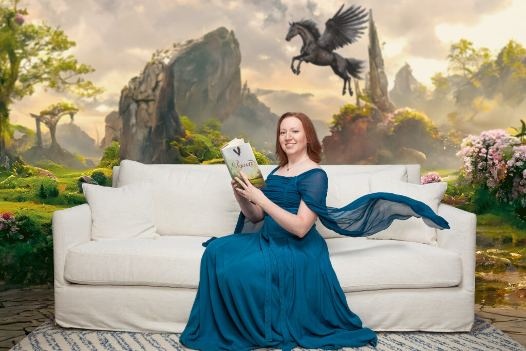 The author, a white woman with red-brown hair, wearing a blue dress with long flowing sleeves, sitting on a white couch holding a hardcover book, with a fantasy background behind her.